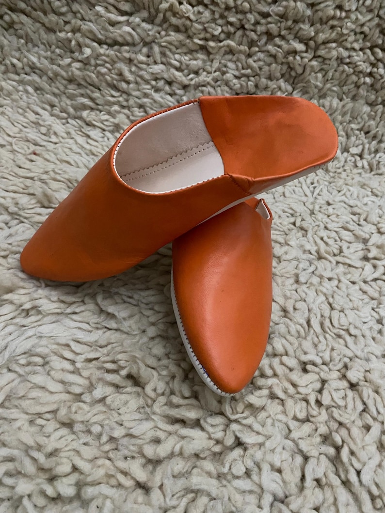 Op de afbeelding: Een paar oranje leren slippers met een puntige neus. De slippers hebben een witte binnenkant en zool. Het schoeisel wordt gepresenteerd op een getextureerd, gebroken wit tapijt.