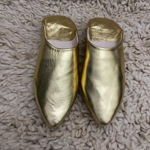 Op de afbeelding: Een paar gouden leren slippers met een puntige neus. De slippers hebben een metallic glans en liggen op een getextureerd, gebroken wit tapijt.