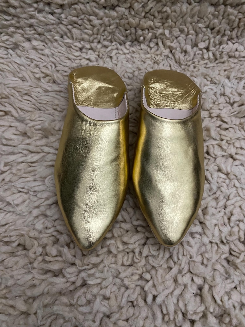 Op de afbeelding: Een paar gouden leren slippers met een puntige neus. De slippers hebben een metallic glans en liggen op een getextureerd, gebroken wit tapijt.