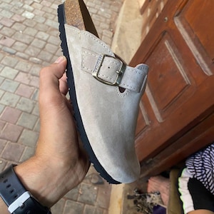 Könnte beinhalten: Hellgrauer Wildleder-Clog mit schwarzer Sohle und Schnallenverschluss. Das Wort "MEDICAL" ist auf der Seite des Schuhs sichtbar. Der Clog hat ein Korkfußbett.