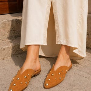 May include: A pair of tan suede mules featuring a pointed toe and a V-shaped cutout at the vamp. The exterior of each shoe is embellished with multiple polished gold-tone metal studs of varying sizes, set against a long cream-colored skirt.