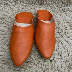 Op de afbeelding: Een paar oranje leren slippers met een puntige neus en een witte binnenkant. De slippers worden getoond op een getextureerd, gebroken wit tapijt.