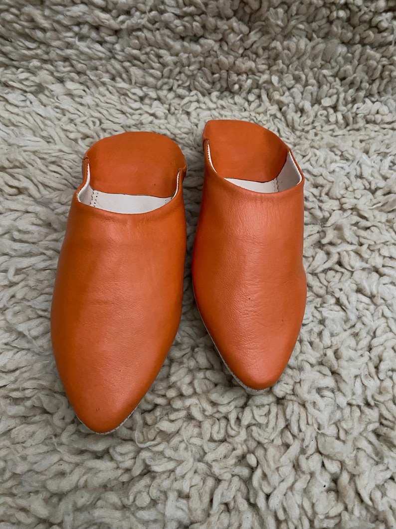 Op de afbeelding: Een paar oranje leren slippers met een puntige neus en een witte binnenkant. De slippers worden getoond op een getextureerd, gebroken wit tapijt.