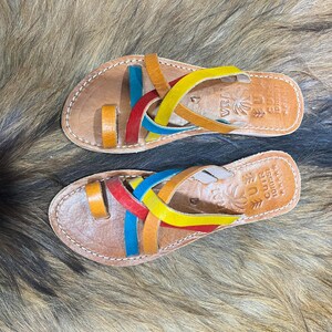 K&ouml;nnte beinhalten: Ein Paar Ledersandalen mit bunten Riemen in Rot, Gelb, Blau und Orange. Die Sandalen haben eine braune Lederbasis und ein Zehenschlaufendesign. Die Sandalen sind handgefertigt und haben N&auml;hte an den R&auml;ndern.