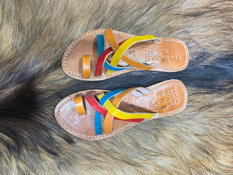 K&ouml;nnte beinhalten: Ein Paar Ledersandalen mit bunten Riemen in Rot, Gelb, Blau und Orange. Die Sandalen haben eine braune Lederbasis und ein Zehenschlaufendesign. Die Sandalen sind handgefertigt und haben N&auml;hte an den R&auml;ndern.