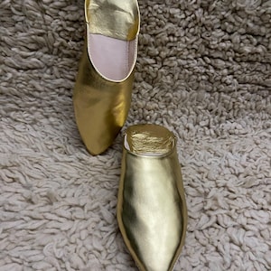 Op de afbeelding: Een paar goudkleurige leren slippers met een spitse neus en een lage hak. De slippers worden getoond op een getextureerd, gebroken wit tapijt. De slippers hebben een metallic glans.