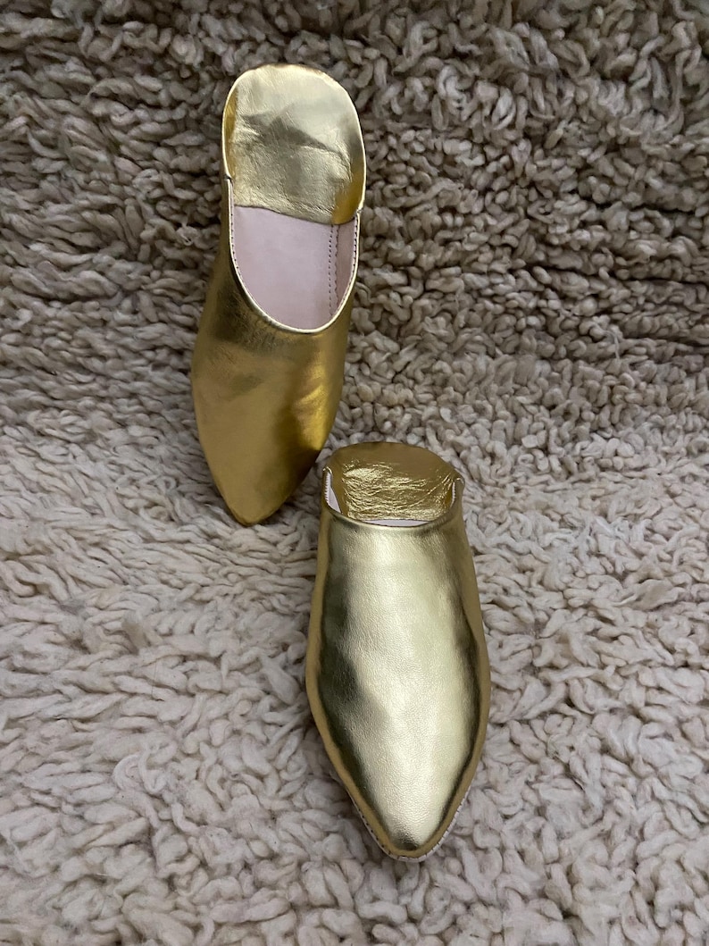 Op de afbeelding: Een paar goudkleurige leren slippers met een spitse neus en een lage hak. De slippers worden getoond op een getextureerd, gebroken wit tapijt. De slippers hebben een metallic glans.