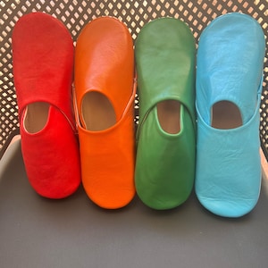 May include: Four pairs of leather slippers in different colors: red, orange, green, and blue. The slippers are lined up in a row on a gray surface.