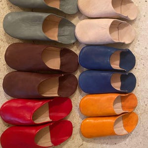 Puede incluir: Un conjunto de diez zapatillas de cuero en varios colores, incluyendo gris, beige, marrón, azul y naranja. Las zapatillas están dispuestas en pares, con los dedos apuntando hacia el lado derecho de la imagen.