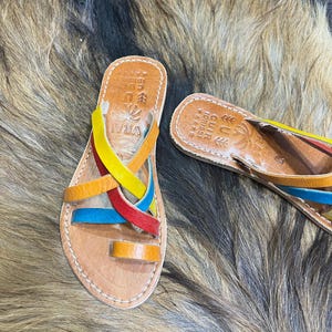 K&ouml;nnte beinhalten: Handgefertigte Ledersandalen mit bunten Riemen in Gelb, Rot, Blau und Orange. Die Sandalen haben ein Naturlederfu&szlig;bett und eine Zehenschlaufe. Die Sandalen werden auf einer strukturierten, braunen Oberfl&auml;che pr&auml;sentiert.
