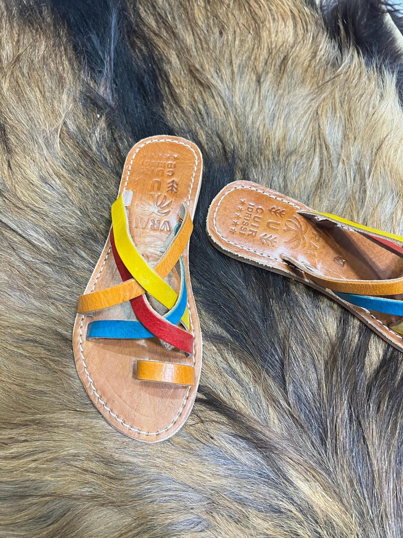 K&ouml;nnte beinhalten: Handgefertigte Ledersandalen mit bunten Riemen in Gelb, Rot, Blau und Orange. Die Sandalen haben ein Naturlederfu&szlig;bett und eine Zehenschlaufe. Die Sandalen werden auf einer strukturierten, braunen Oberfl&auml;che pr&auml;sentiert.