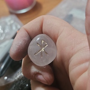 May include: A clear, rounded rose quartz stone with a gold-colored etched symbol. The stone is held in a hand, with a lit pink candle in the background. The symbol is a stylized cross.