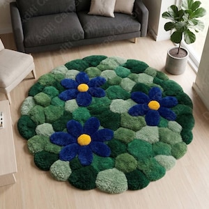 May include: A round, tufted area rug featuring a textured surface of various green shades resembling moss, accented by three large, dark blue flowers with bright yellow centers. The rug has a thick, plush pile and a distinct, organic circular shape.