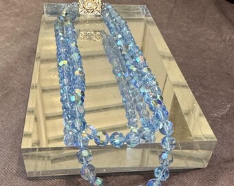 May include: A multi-strand necklace featuring iridescent light blue beads. The necklace is connected by a square, silver-colored clasp. The necklace is displayed on a clear, rectangular surface.