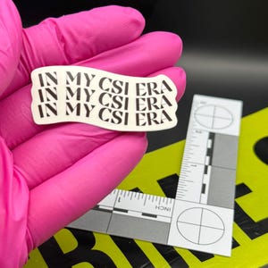 May include: A white sticker with the text "IN MY CSI ERA" in black, held by a gloved hand. The sticker is slightly curved. A crime scene ruler and yellow caution tape are in the background.