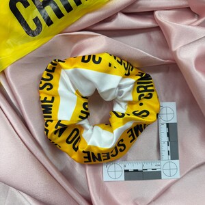 May include: A yellow and white scrunchie with the words "CRIME SCENE DO NOT CROSS" printed on it. The scrunchie is on a pink satin fabric. A yellow "CRIME" tape is in the background.