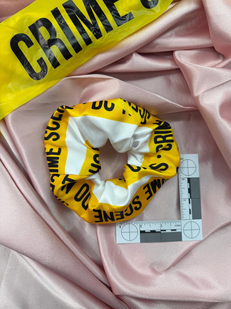 May include: A yellow and white scrunchie with the words "CRIME SCENE DO NOT CROSS" printed on it. The scrunchie is on a pink satin fabric. A yellow "CRIME" tape is in the background.