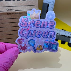 May include: A holographic sticker with the words "Scene Queen" in pink, featuring a crown, fingerprint, magnifying glass, camera, and other crime scene-related icons. The sticker is held by a gloved hand.