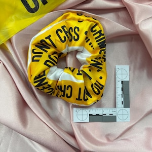May include: Yellow scrunchie with black text reading "DO NOT CROSS CRIME SCENE" and white stripes. A yellow crime scene tape and a forensic ruler are also in the image. The scrunchie is on a pink satin surface.