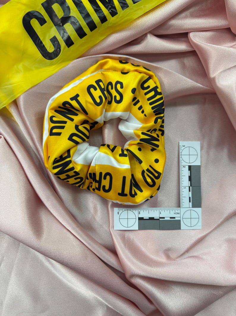 May include: Yellow scrunchie with black text reading "DO NOT CROSS CRIME SCENE" and white stripes. A yellow crime scene tape and a forensic ruler are also in the image. The scrunchie is on a pink satin surface.