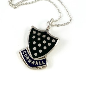 May include: Silver necklace with a shield pendant. The pendant is black with white dots and a blue banner that reads "Cornwall". The necklace is on a white background.