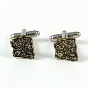 May include: A pair of silver cufflinks with a gold-toned outline of the state of Arizona. The cufflinks feature the words "PHOENIX" and "TUCSON" with a star in between.
