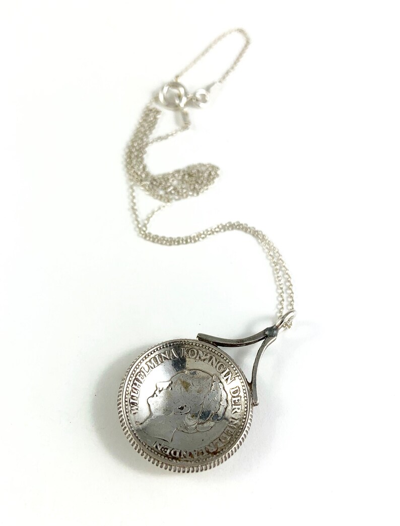 May include: Silver necklace with a coin pendant featuring a profile of Wilhelmina, Queen of the Netherlands. The coin is attached to a silver chain with a clasp.