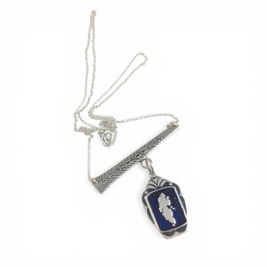 May include: A silver necklace with a bar pendant and a rectangular pendant. The bar pendant has etched designs. The rectangular pendant has a blue background with a white shape in the center.