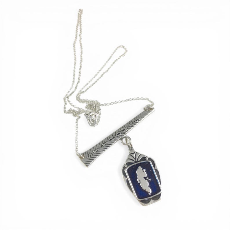 May include: A silver necklace with a bar pendant and a rectangular pendant. The bar pendant has etched designs. The rectangular pendant has a blue background with a white shape in the center.