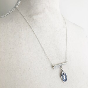 May include: A silver necklace with a rectangular pendant featuring a blue and white design. The pendant hangs from a bar with a leaf design, suspended from a delicate chain. The necklace is displayed on a mannequin.