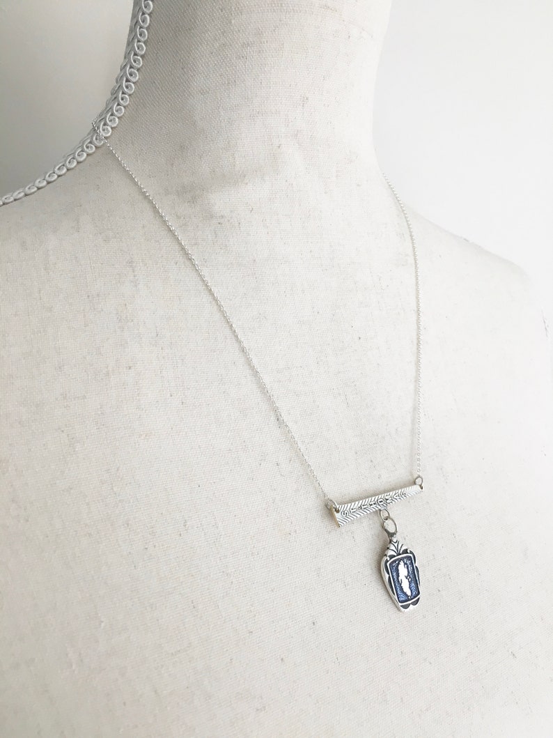 May include: A silver necklace with a rectangular pendant featuring a blue and white design. The pendant hangs from a bar with a leaf design, suspended from a delicate chain. The necklace is displayed on a mannequin.