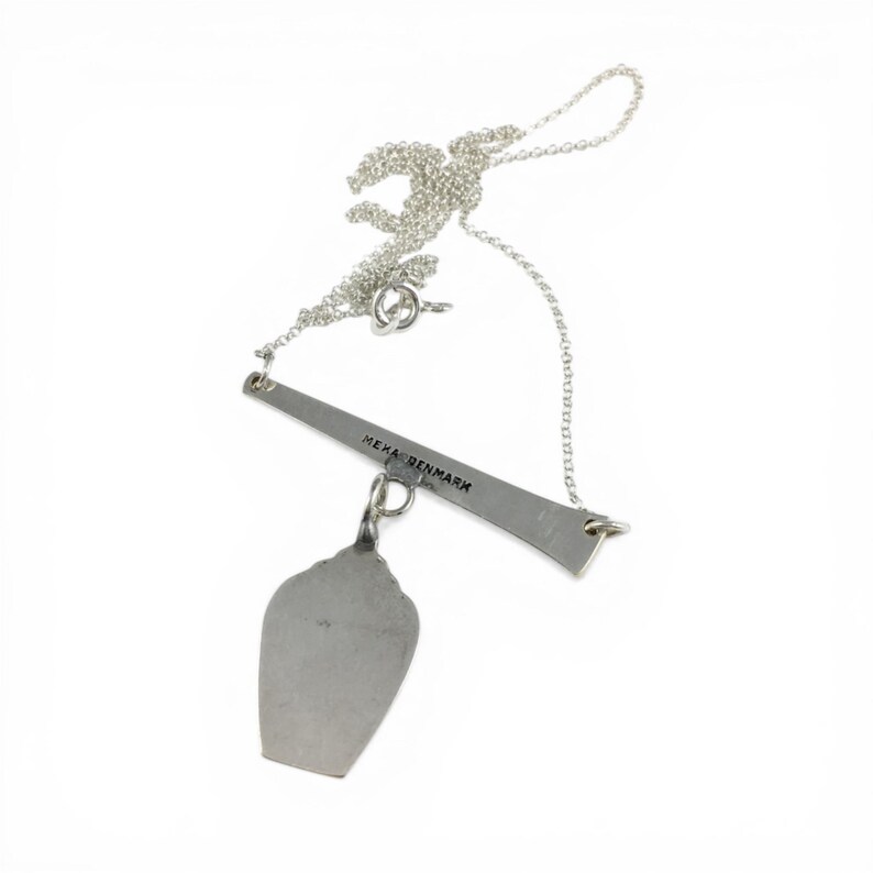 May include: A silver necklace with a pendant. The pendant is a stylized, irregular shape suspended from a horizontal bar. The bar has the text "MEHA DENMARK" engraved on it. The necklace chain is delicate and silver.