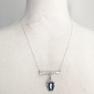 May include: Silver-toned necklace featuring a rectangular pendant with a blue background and a white silhouette. The pendant hangs from a decorative bar with a textured design. The necklace is displayed on a mannequin.