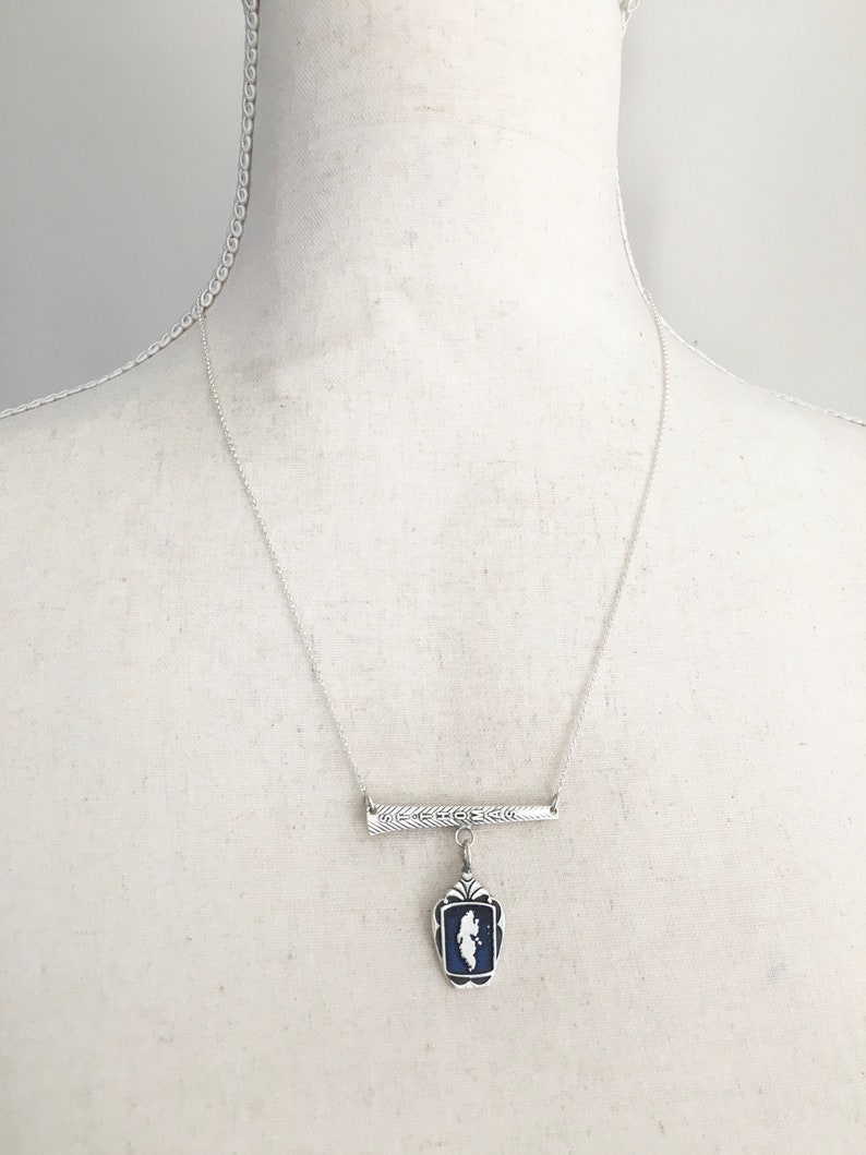 May include: Silver-toned necklace featuring a rectangular pendant with a blue background and a white silhouette. The pendant hangs from a decorative bar with a textured design. The necklace is displayed on a mannequin.