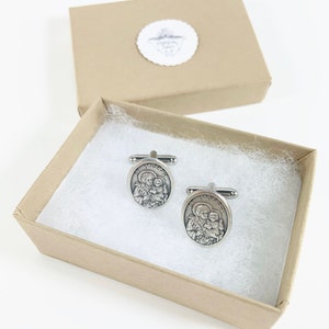 May include: A pair of silver cufflinks with a religious image of Saint Joseph and the baby Jesus. The cufflinks are in a small, brown gift box with white tissue paper.