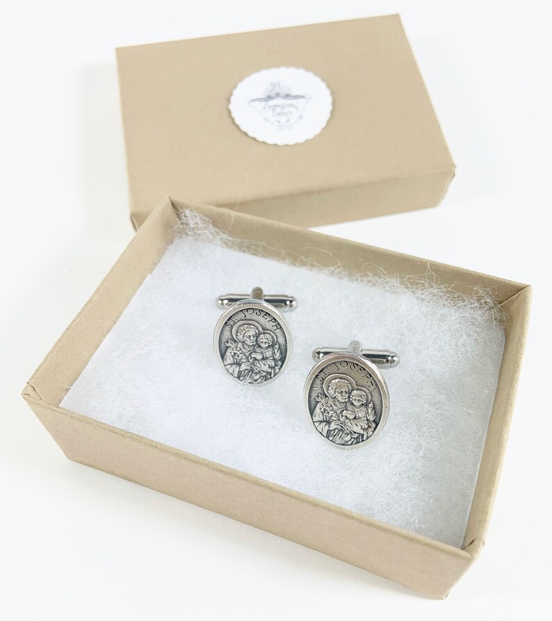 May include: A pair of silver cufflinks with a religious image of Saint Joseph and the baby Jesus. The cufflinks are in a small, brown gift box with white tissue paper.