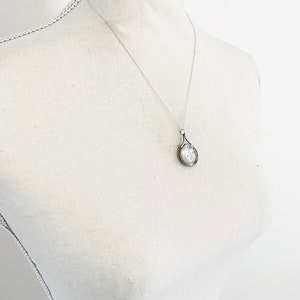 May include: A silver chain necklace with a silver pendant shaped like a teardrop. The pendant has a textured surface and a small, clear stone in the center.