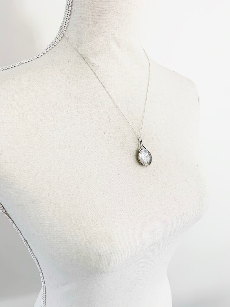 May include: A silver chain necklace with a silver pendant shaped like a teardrop. The pendant has a textured surface and a small, clear stone in the center.