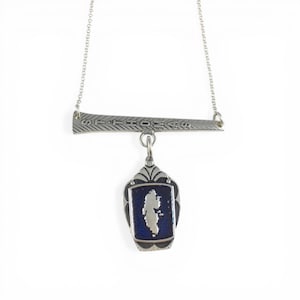 May include: Silver-toned necklace featuring a horizontal bar with the inscription "ST THOMAS" and a pendant. The pendant has a dark blue background with a white island shape. The necklace is suspended by a delicate chain.