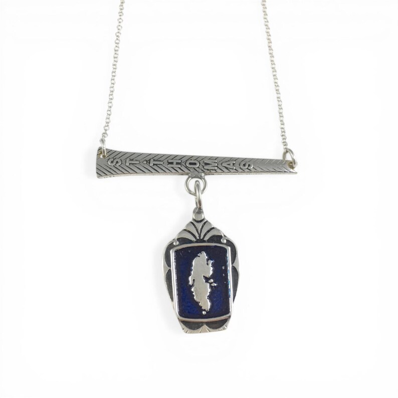 May include: Silver-toned necklace featuring a horizontal bar with the inscription "ST THOMAS" and a pendant. The pendant has a dark blue background with a white island shape. The necklace is suspended by a delicate chain.
