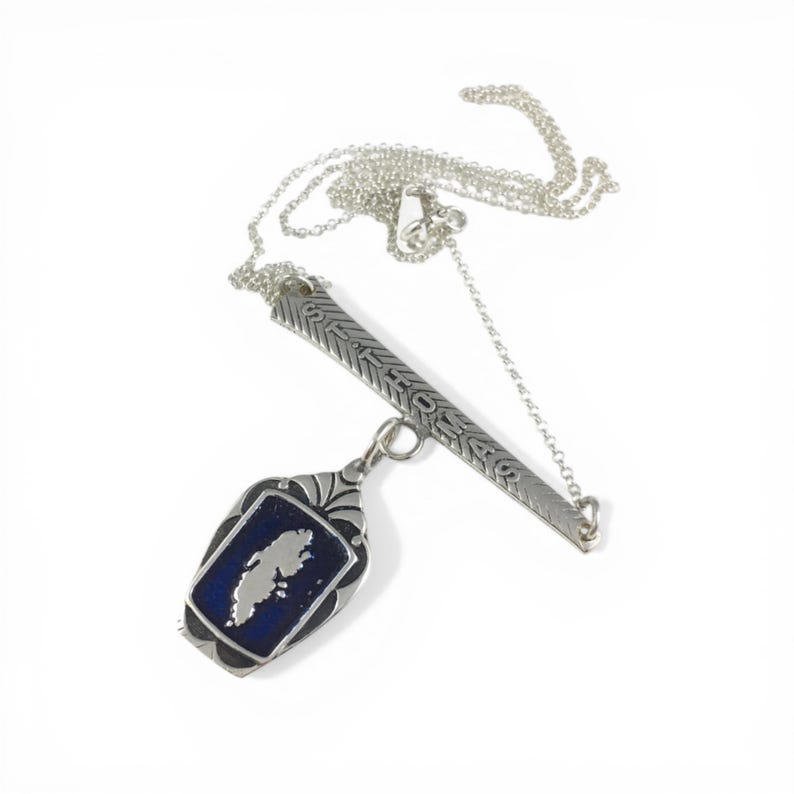 May include: Silver-toned necklace with a rectangular pendant and a blue enamel background. The pendant features a white island outline. The necklace also includes a decorative bar with etched details.