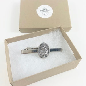 May include: A silver tie clip with a religious image of Saint Joseph holding the baby Jesus. The tie clip is in a small, brown gift box with white lining.