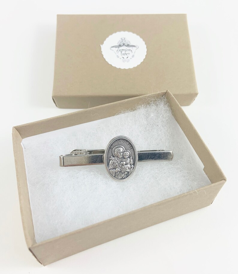 May include: A silver tie clip with a religious image of Saint Joseph holding the baby Jesus. The tie clip is in a small, brown gift box with white lining.