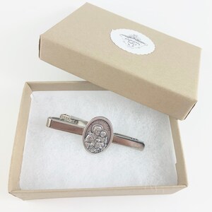 May include: A silver tie clip with a round, oval-shaped charm featuring a depiction of Saint Joseph holding the baby Jesus. The tie clip is in a small, white, rectangular box with a brown lid.