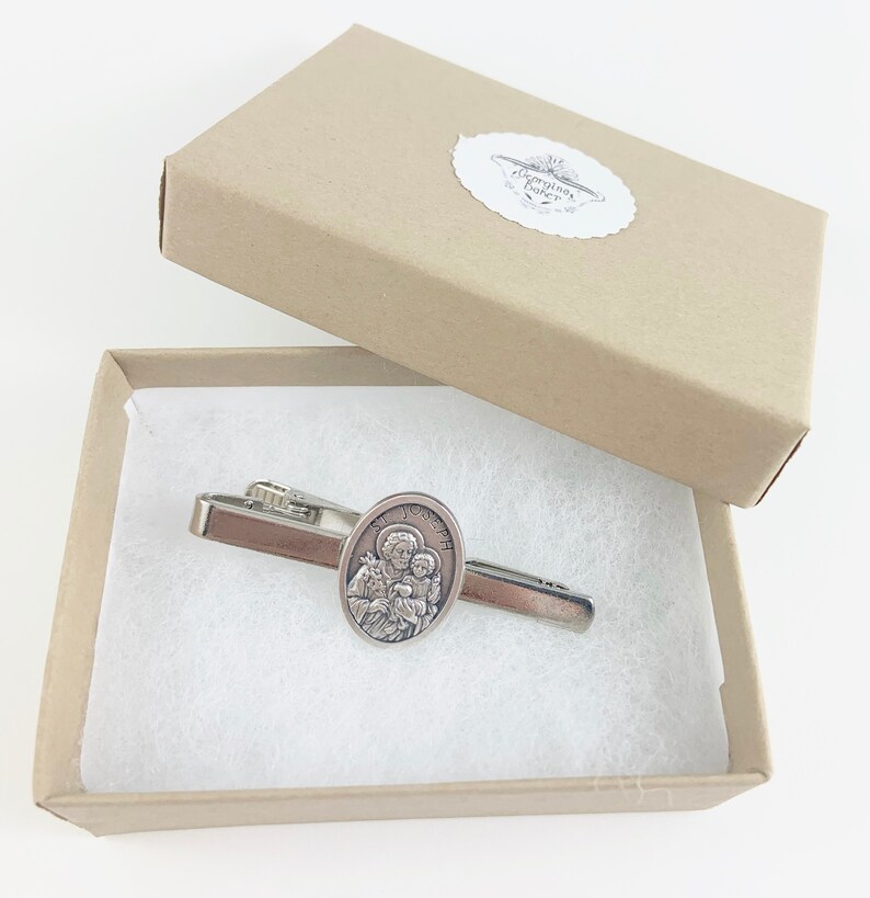 May include: A silver tie clip with a round, oval-shaped charm featuring a depiction of Saint Joseph holding the baby Jesus. The tie clip is in a small, white, rectangular box with a brown lid.