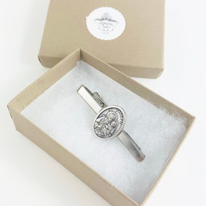 May include: A silver tie clip with a round, silver charm featuring a depiction of Saint Joseph. The charm has the words "St. Joseph" inscribed around the edge. The tie clip is in a small, brown box with white tissue paper.
