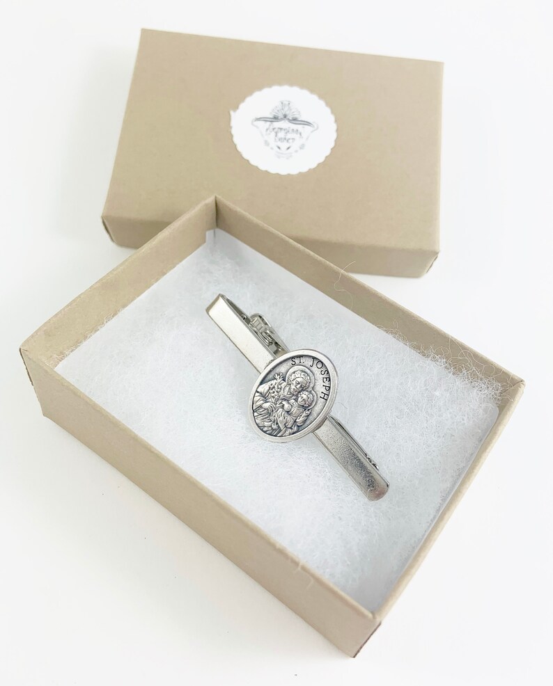 May include: A silver tie clip with a round, silver charm featuring a depiction of Saint Joseph. The charm has the words "St. Joseph" inscribed around the edge. The tie clip is in a small, brown box with white tissue paper.