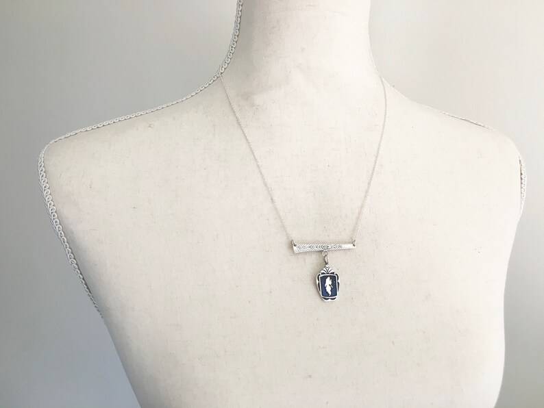 May include: A silver-toned necklace with a bar pendant and a rectangular blue and white pendant. The bar pendant has engraved text. The necklace is displayed on a mannequin.