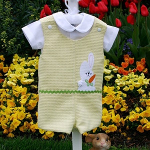 Op de afbeelding: Een gele en groene baby romper met een witte konijn en wortel applicatie. De romper heeft een witte kraag en hangt aan een witte standaard.