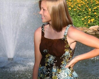 May include: A young woman wearing a brown and blue floral sundress with a brown bodice and a white and gold bottom. She is standing in front of a fountain.